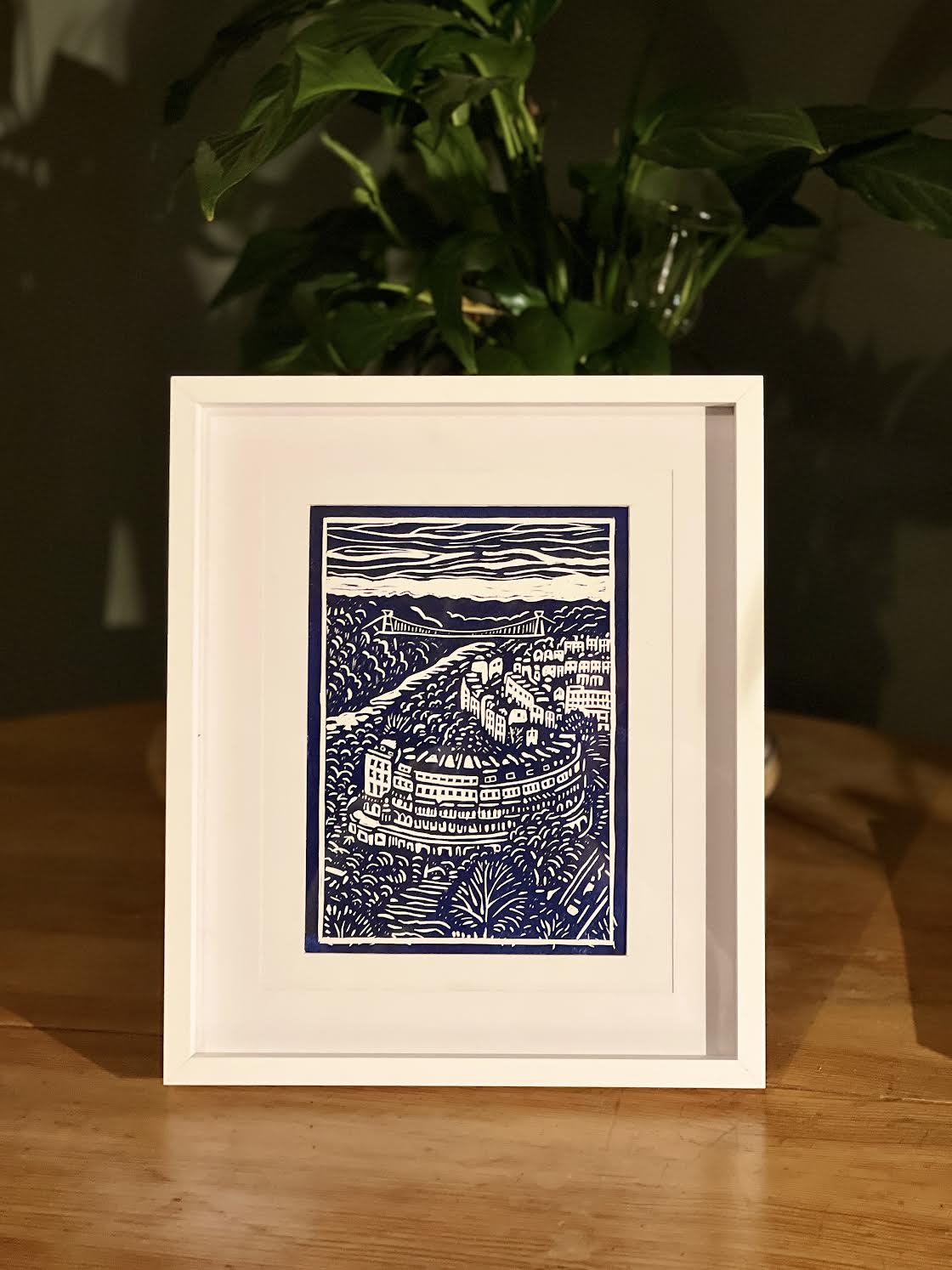 Framed artwork of a blue and white landscape on a wooden surface with a plant in the background.