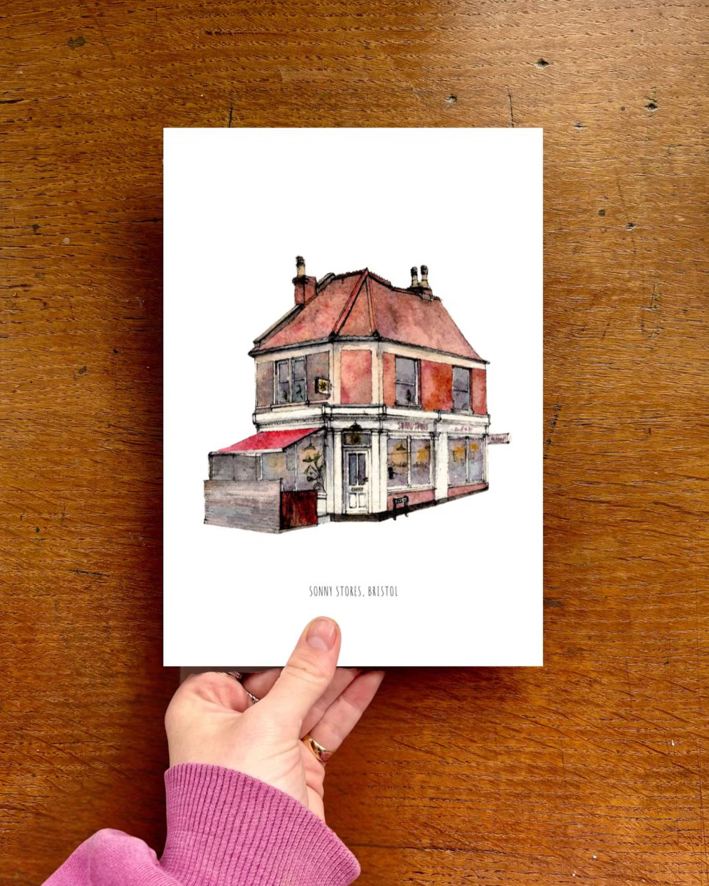 Hand holding a print of a house illustration on a wooden surface