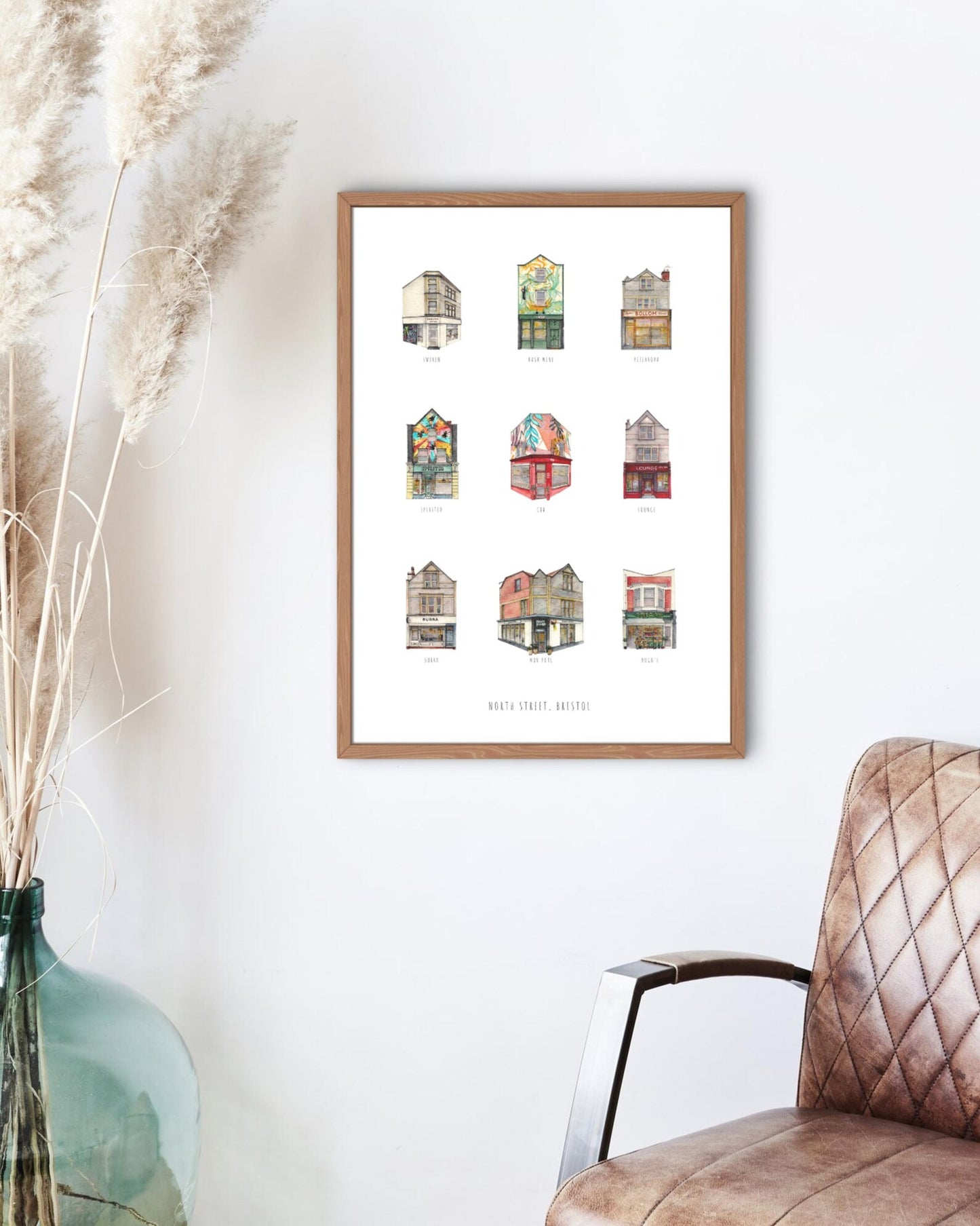 Framed artwork of colorful building illustrations on a white wall with a chair and decorative vase.