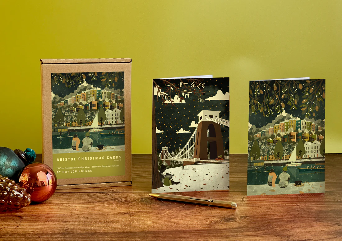 Three Christmas cards with a cityscape design on a wooden surface against a yellow wall.
