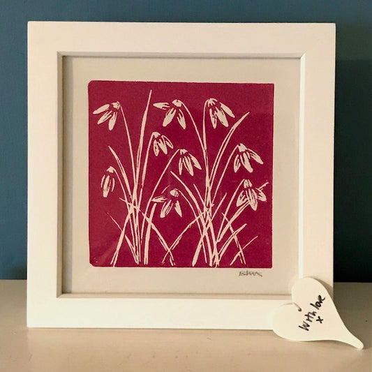 Pink background with snowdrops lino print in a white frame.