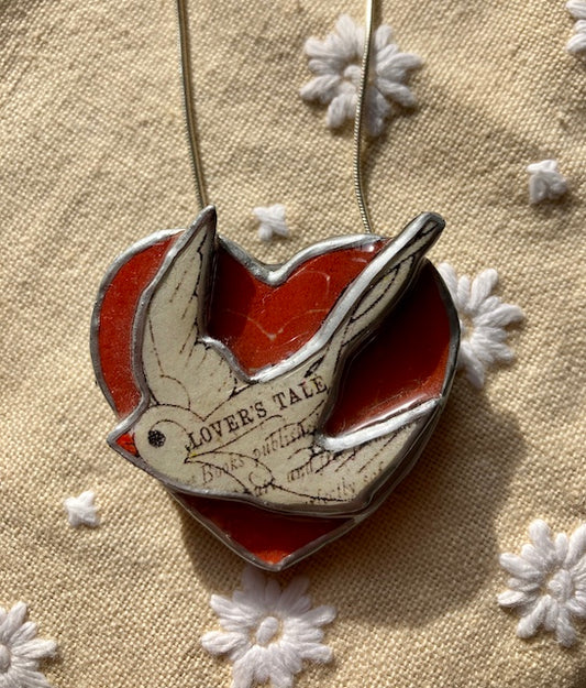 This pendant is made by Ellymental a Cardiff based Artist.  by layering vintage paper and metal and resin.  A white dove protrudes from a red heart.  The text Lovers Tale across its body.