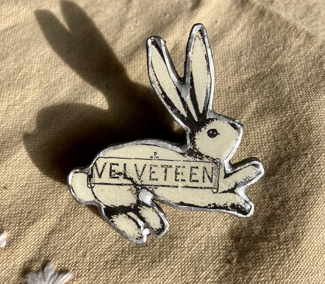 A white rabbit brooch with the text Velveteen across its body.  Handmade from recycled materials of layered paper, metal and resin.  Made by Ellymental a Cardiff based artist.