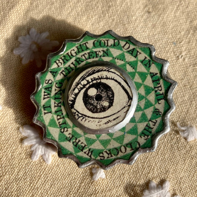 This George Orwell Eye brooch is handmade by Elemental a Cardiff artist.  Made from layered paper and metal a laquered with resin to seal.  The text around the eye reads It was a bright cold day in April and the clocks were striking the thirteen.