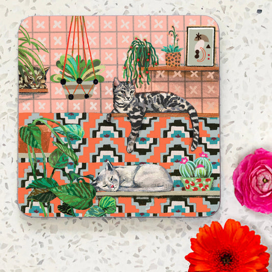 Coaster with two cats lying on shelves with patterned tiles and plants. Coral pink background.