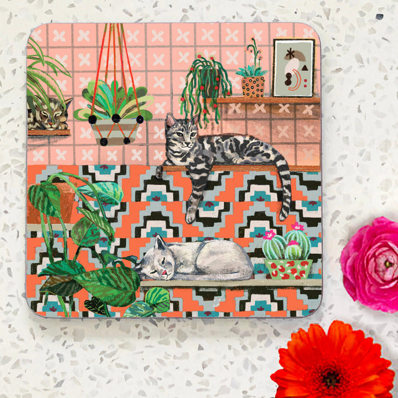 Coaster with two cats lying on shelves with patterned tiles and plants. Coral pink background.