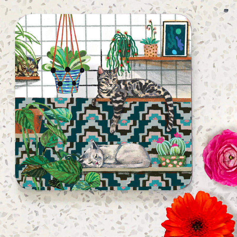 Coaster with two cats lying on shelves with patterned tiles and plants. White background.
