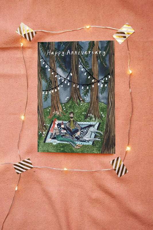 watercolour greetings card with couple and dog picnicing in a woods with lights in the trees and the words ''happy anniversary''