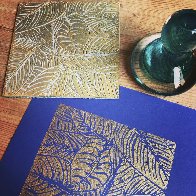 Trelawney Designs hand cut lino print with water based ink. Botanical leaves in Gold on a Dark blue back ground. Framed in a white frame.  Edit alt text