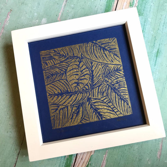 Trelawney Designs hand cut lino print with water based ink.  Botanical leaves in Gold on a Dark blue back ground.  Framed in a white frame.