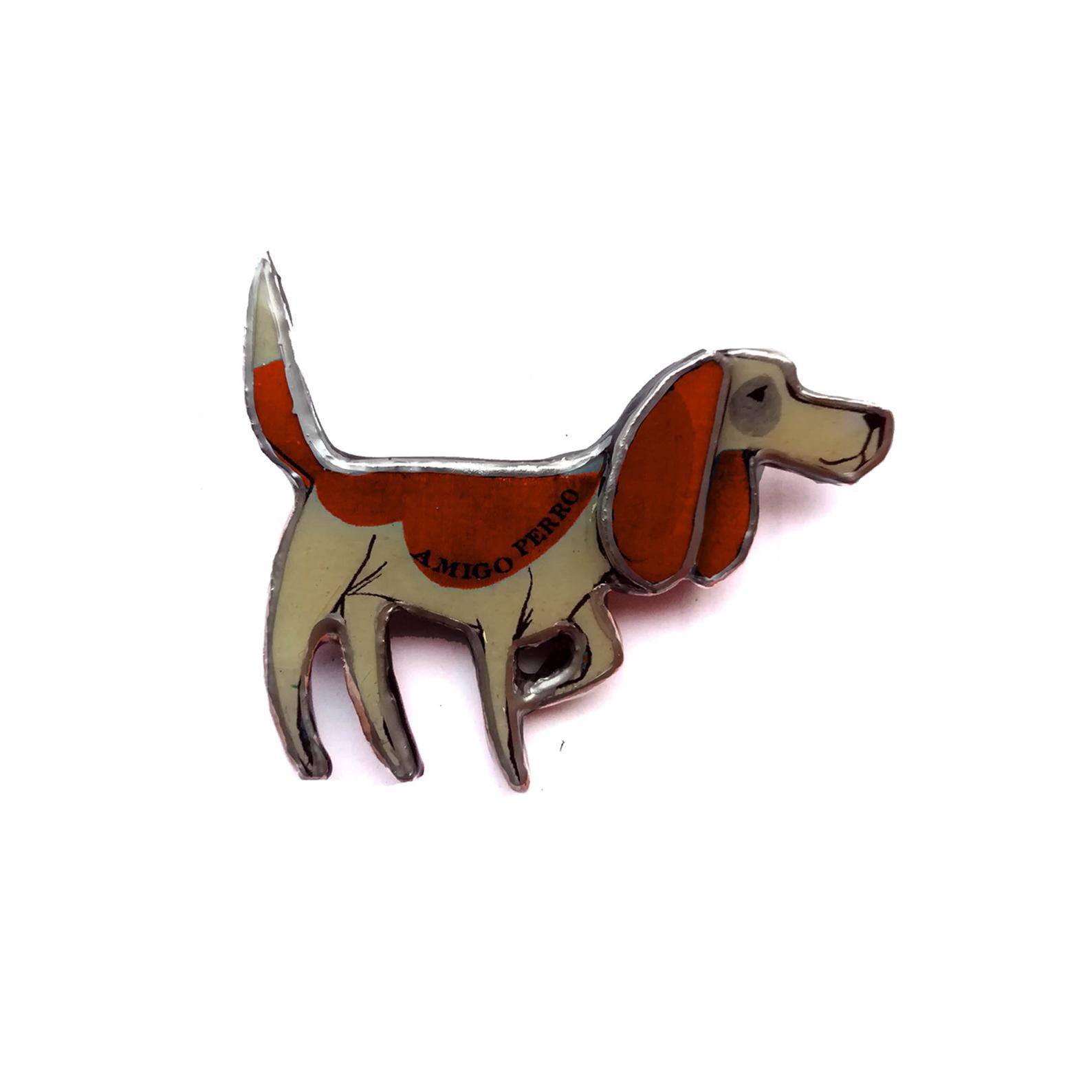 A beagle with the words amigo per on its body.  Handmade brooch made from layered paper, metal and resin.